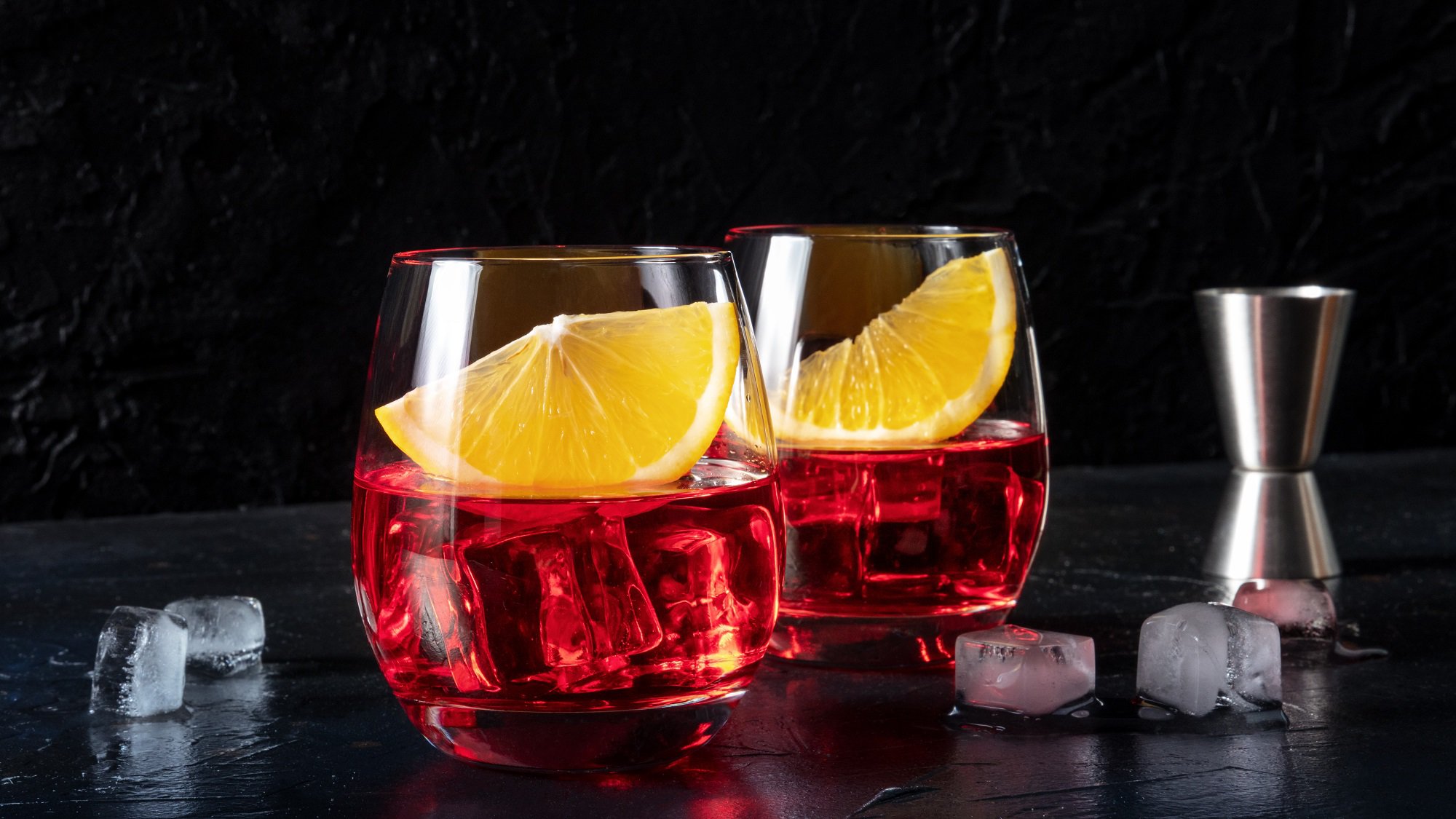 Zwei Gläser Wermut-Spritz mit Eiswürfeln und je einer Orangenspalte vor dunklem Hintergrund, daneben ein Cocktail-Messbecher.