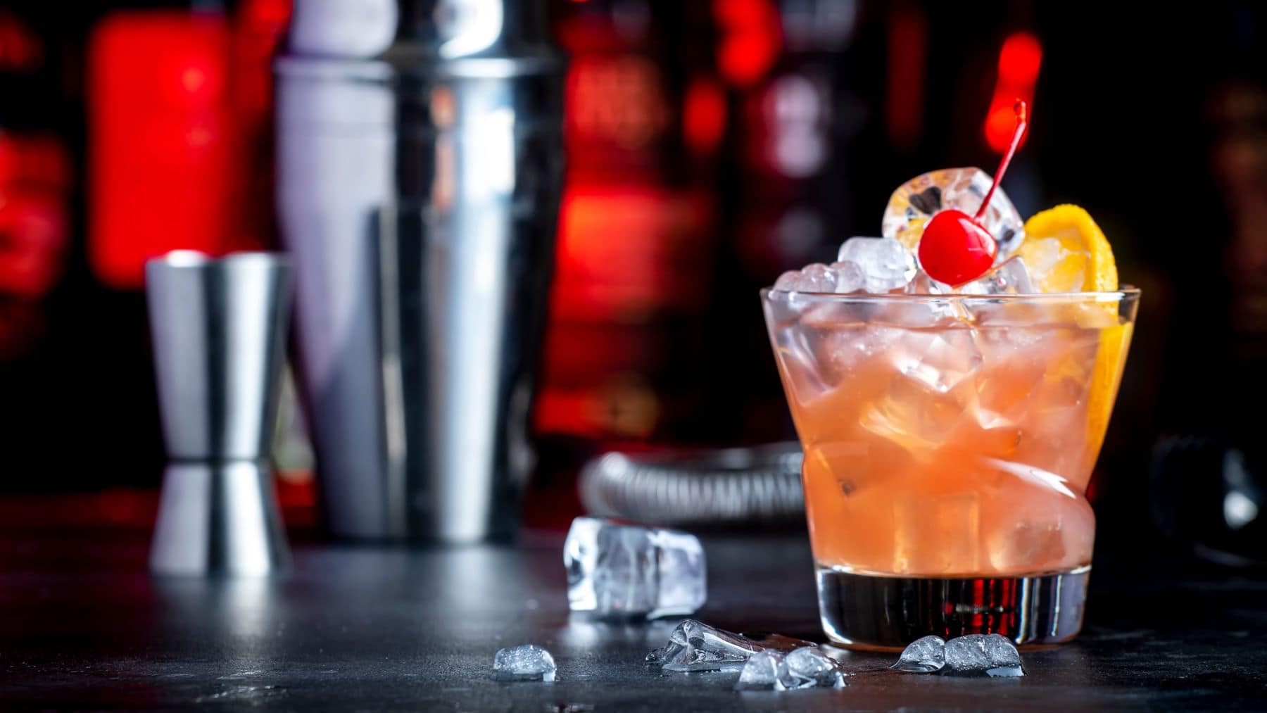 Whiskey Daisy mit Zitronenscheibe, Cocktail-Kirsche und Eis im Glas auf dunklem Untergrund. Im Hintergrund eine unscharfe Bar, Barmaß, Cccktail-Shaker und Eiswürfel. Frontalansicht.