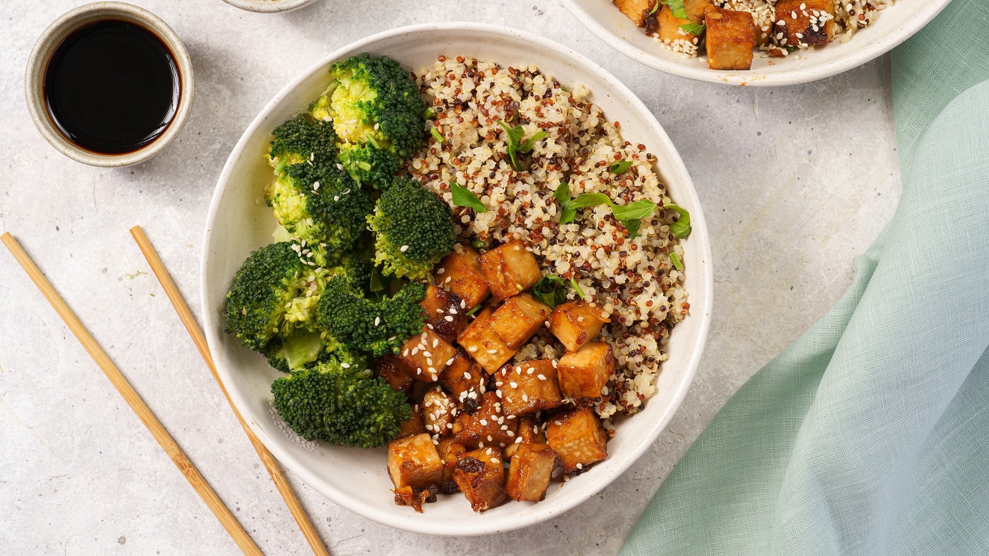 Eine Brokkoli-Quinoa-Bowl mit Tofu in der Draufsicht, daneben 2 Essstäbchen und eine Schale Sojasauce.