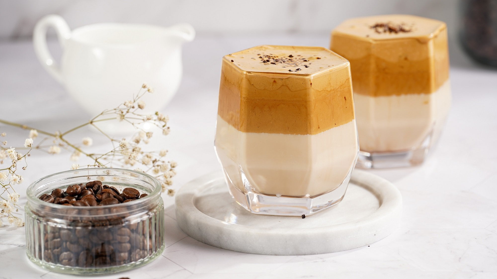 2 Gläser Dalgona Pumpkin Spice Latte, eins davon auf einem weißen Marmor-Untersetzer, daneben eine Schale mit Kaffeebohnen und ein Milchkännchen.