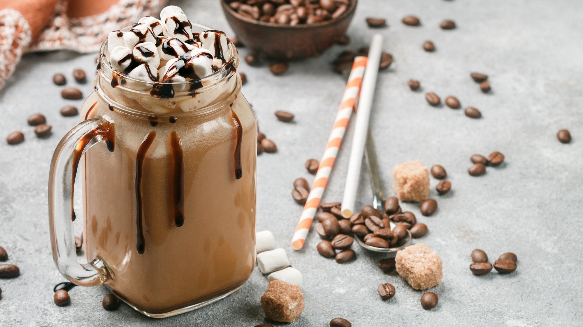 Kaffee-Shake in einem Glas mit Henkel, getoppft mit Marshmallows und Schokosauce, daneben Kaffeebohnen und Stohhalme.