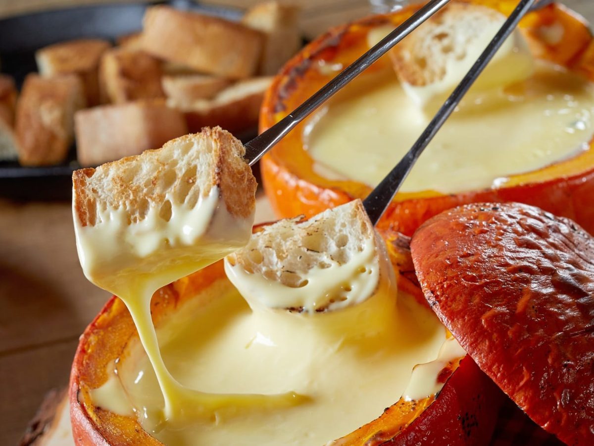 Zwei Kürbisse mit Käse-Fondue auf einem Holztisch seitlich fotografiert. Daneben noch eine gusseiserne Pfanne mit Brotstückchen.