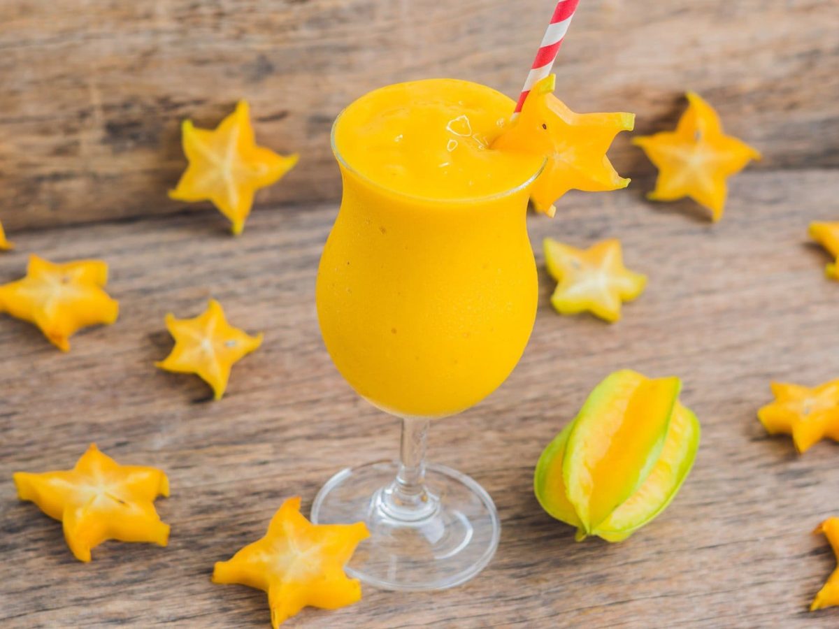 Draufsicht, schräg: ein Sternfrucht-Mango-Smoothie im Glas mit einem Strohhalm. Drum herum liegen aufgeschnittene Sternfrucht-Scheiben.
