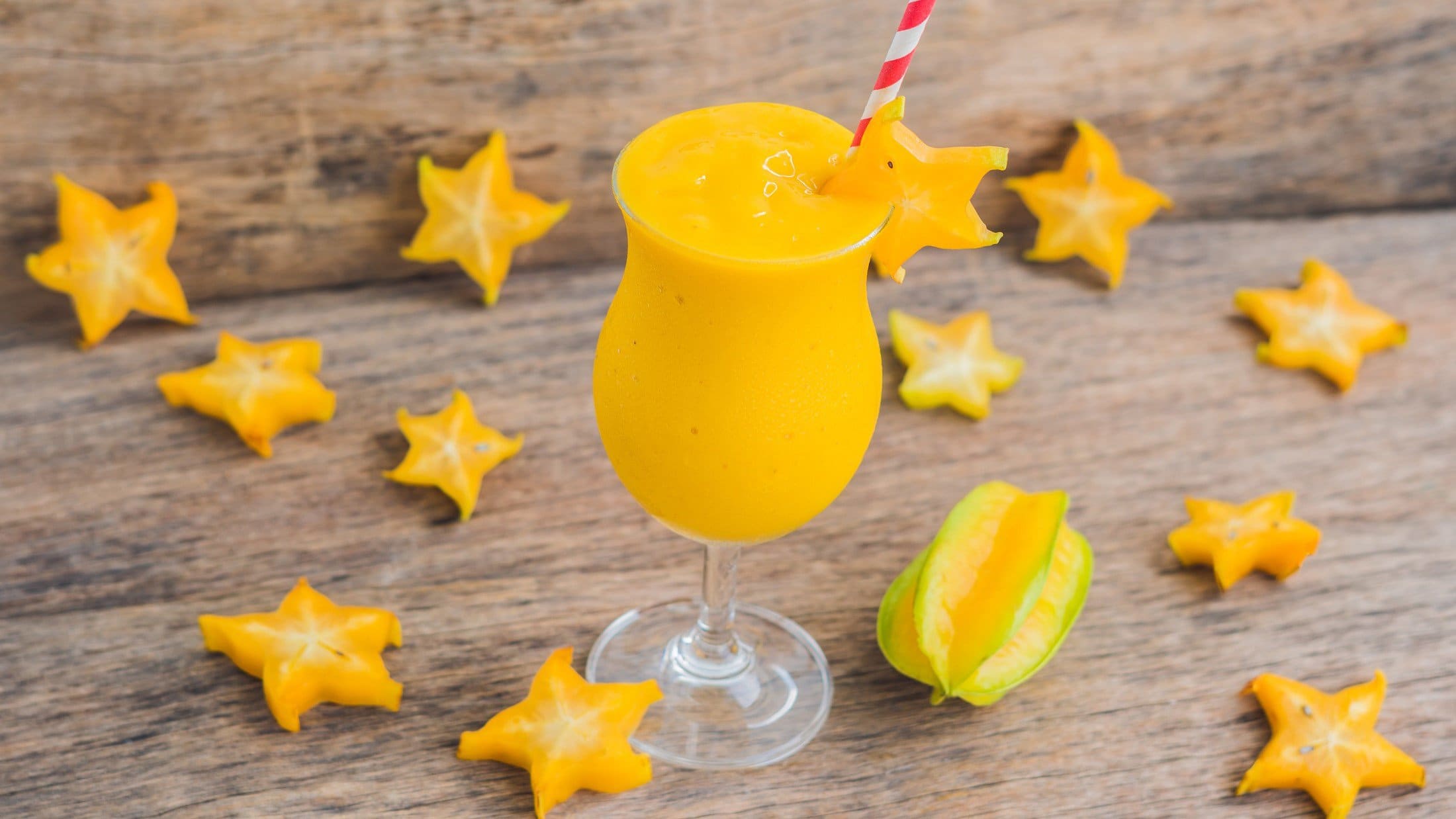 Draufsicht, schräg: ein Sternfrucht-Mango-Smoothie im Glas mit einem Strohhalm. Drum herum liegen aufgeschnittene Sternfrucht-Scheiben.
