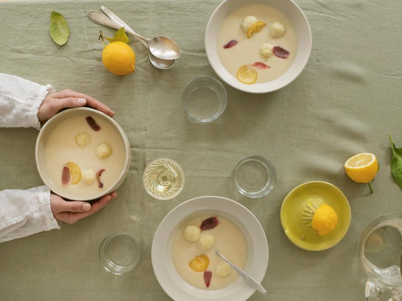3 Schüssel Zitronensuppe mit Klößen aus dem Kochbuch "Suppenkult", nach einer Schüssel greifen zwei Hände, drumherum verteilt Zitronen, Gläser und Löffel, Draufsicht.