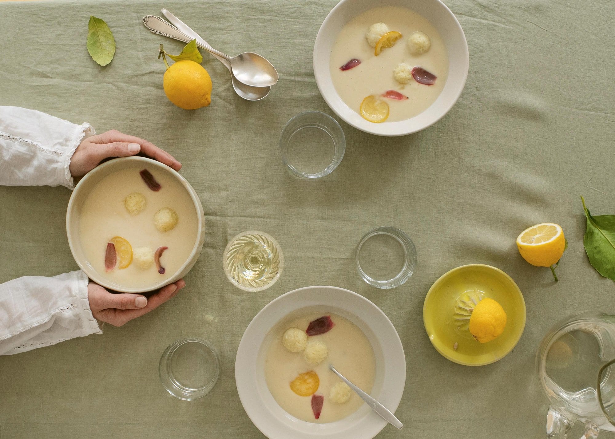 3 Schüssel Zitronensuppe mit Klößen aus dem Kochbuch "Suppenkult", nach einer Schüssel greifen zwei Hände, drumherum verteilt Zitronen, Gläser und Löffel, Draufsicht.