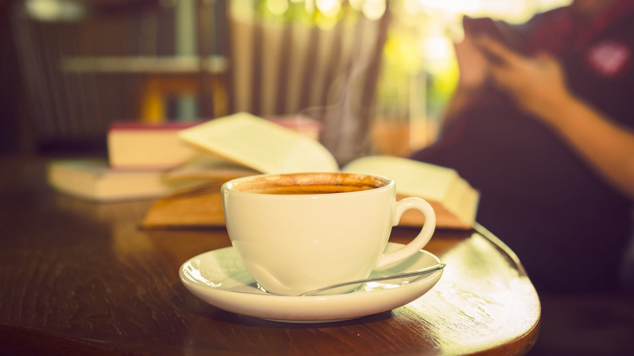 Frontal eine weiße Kaffeetasse mit Untertasse steht in einem Raum auf einem dunklen, runden Holztischchen. Dahinter liegen mehrere Bücher, zum teil aufgeschlagen.