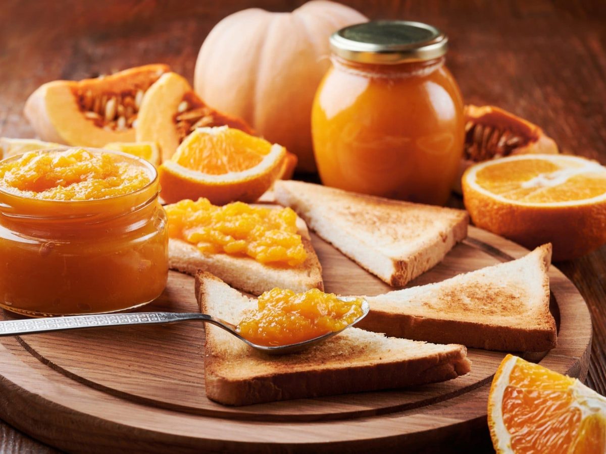 Draufsicht, schräg: Zwei Gläser Kürbis-Orangen-Aufstrich sowie Toastscheiben, Kürbisfleisch und Orangenhälften liegen auf einem runden Holzbrett.