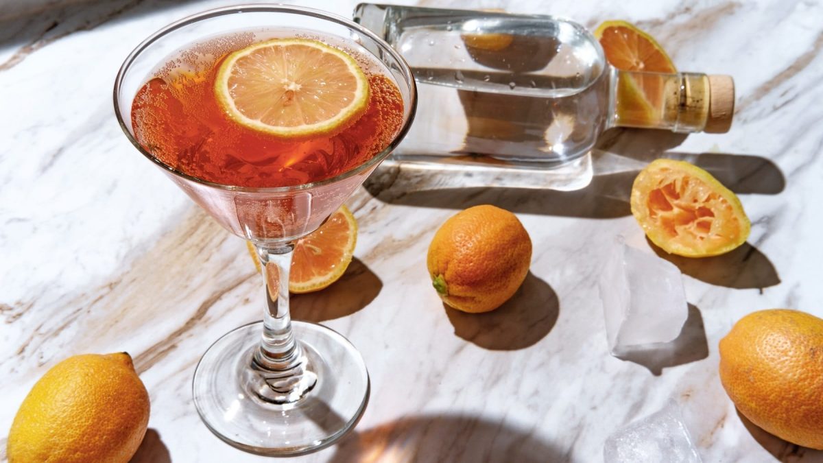 Ein Martini-Glas mit dem Pink Lemon Drop auf einem Marmortisch mit Zitronen, Grapefruit und einer Glasflasche mit Wodka. Alles von schräg oben fotografiert.
