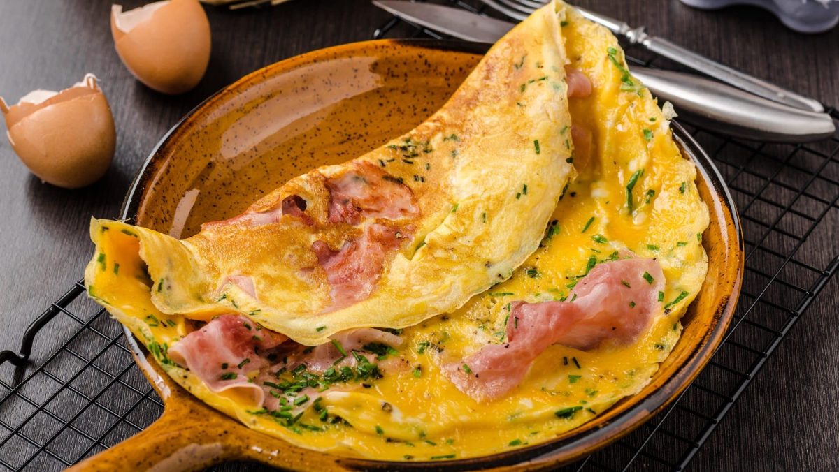 Draufsicht, schräg: Ein italienische Frittata al Prosciuttoin einer Pfanne auf gebranntem und glasiertem Ton. Daneben liegen Besteck und zwei aufgeschlagene Eier.