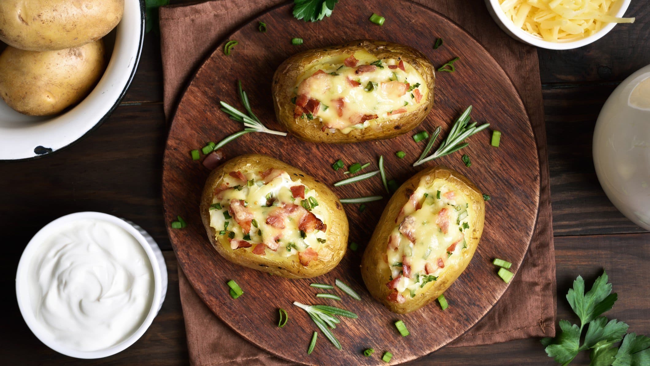 Carbonara-Kartoffeln, gefüllt mit Speck und Käse auf runden Holzplatte. Daneben die einzelnen Zutaten.