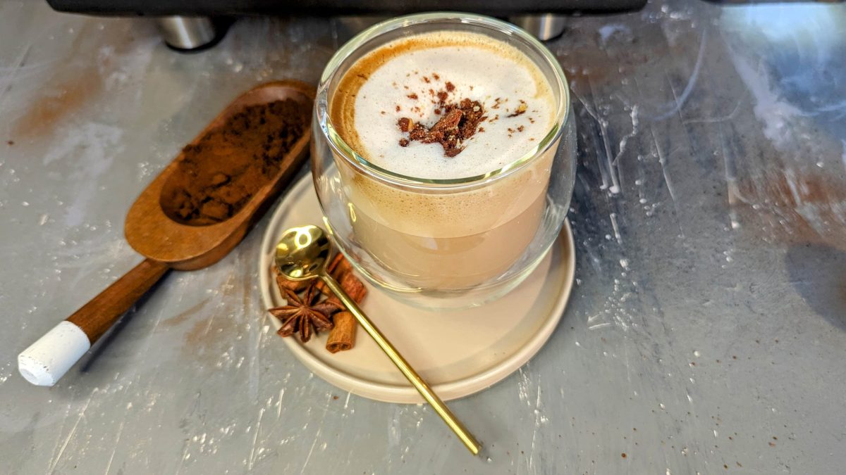 Ein Glas mit dem Kokos-Lebkuchen-Latte auf einem grauen Steintisch mit winterlichen Gewürzen und Kaffeepulver.
