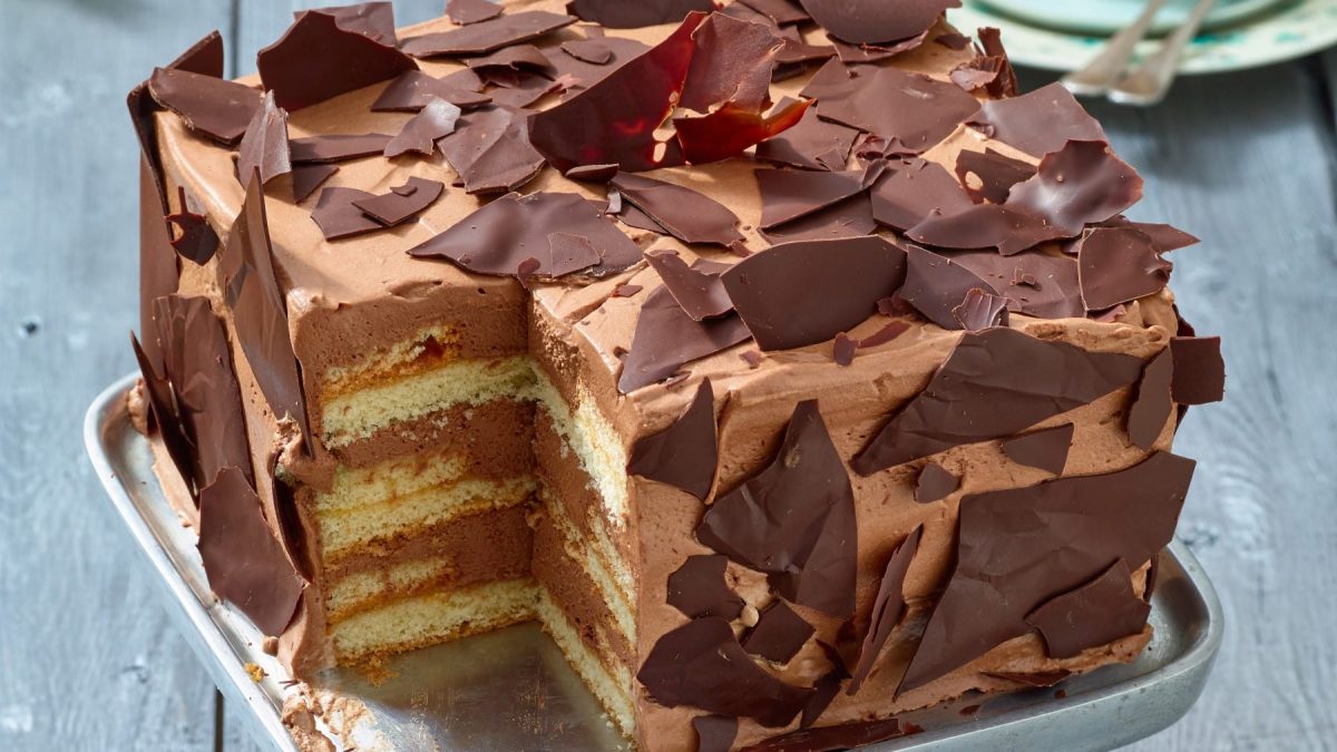 Angeschnittene, viereckige Schoko-Sahne-Torte mit mehreren Schichten und Schokosplittern verziert vor grauem Hintergrund.