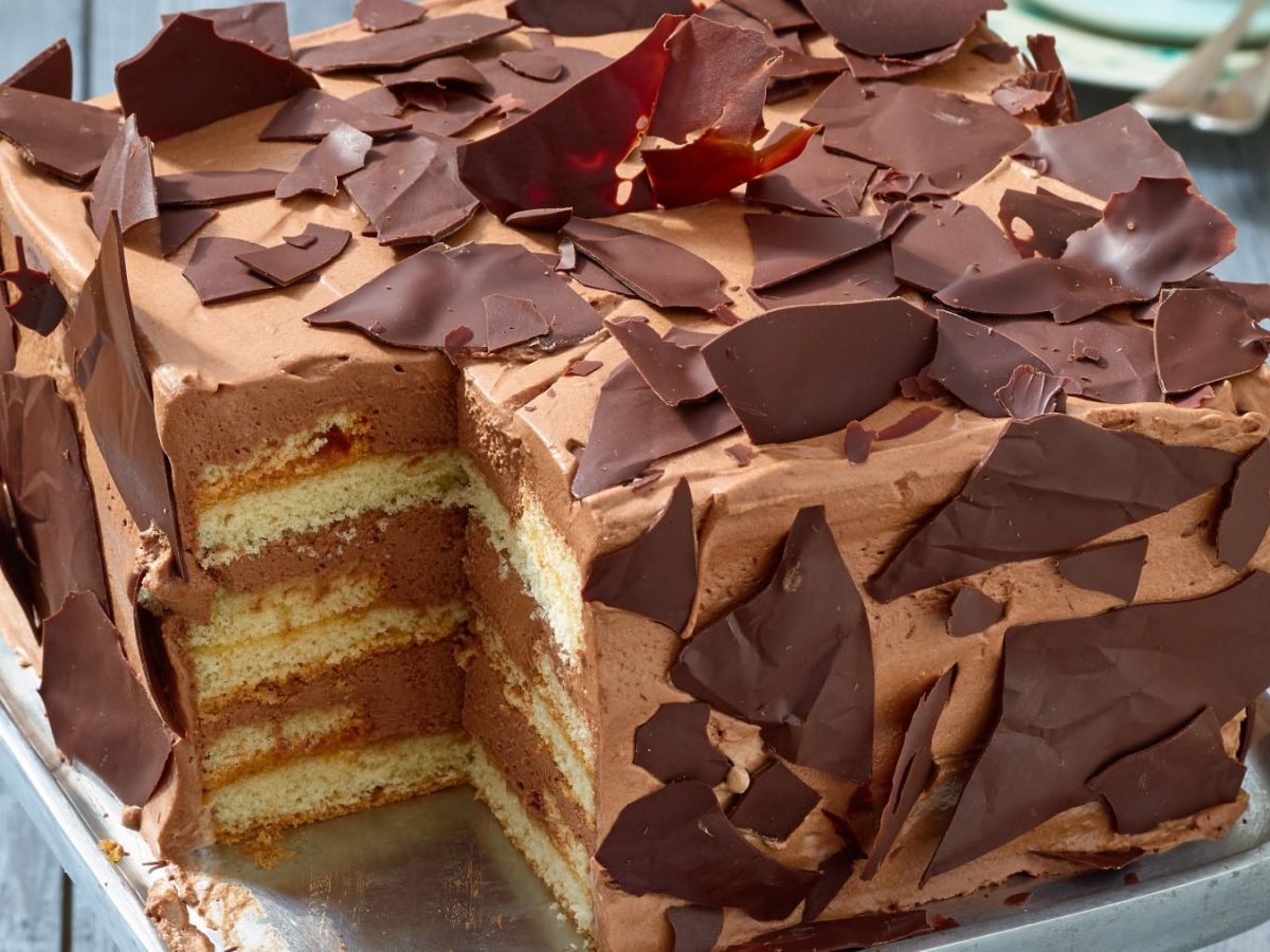Angeschnittene, viereckige Schoko-Sahne-Torte mit mehreren Schichten und Schokosplittern verziert vor grauem Hintergrund.