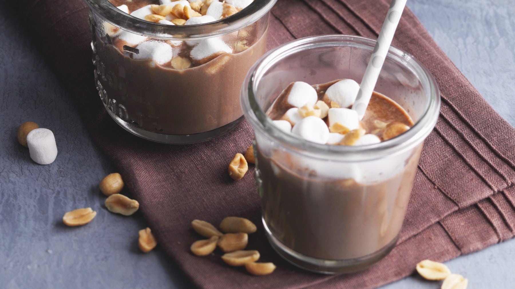 Süß-salzige heiße Schokolade in zwei Gläsern mit Marshmallows und Erdnüssen auf braunem Stoff, grauer Untergrund.