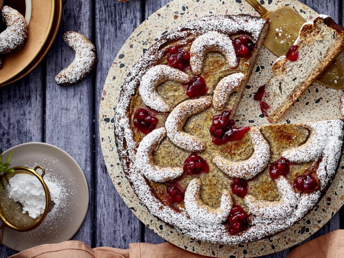 Ein Vanillekipferl-Kuchen auf einem Mosaik-Servierteller auf einem hölzernen Hintergrund. Links ein Teller mit Vanillekipferl und links unten ein kleinen Sieb mit Puderzucker.