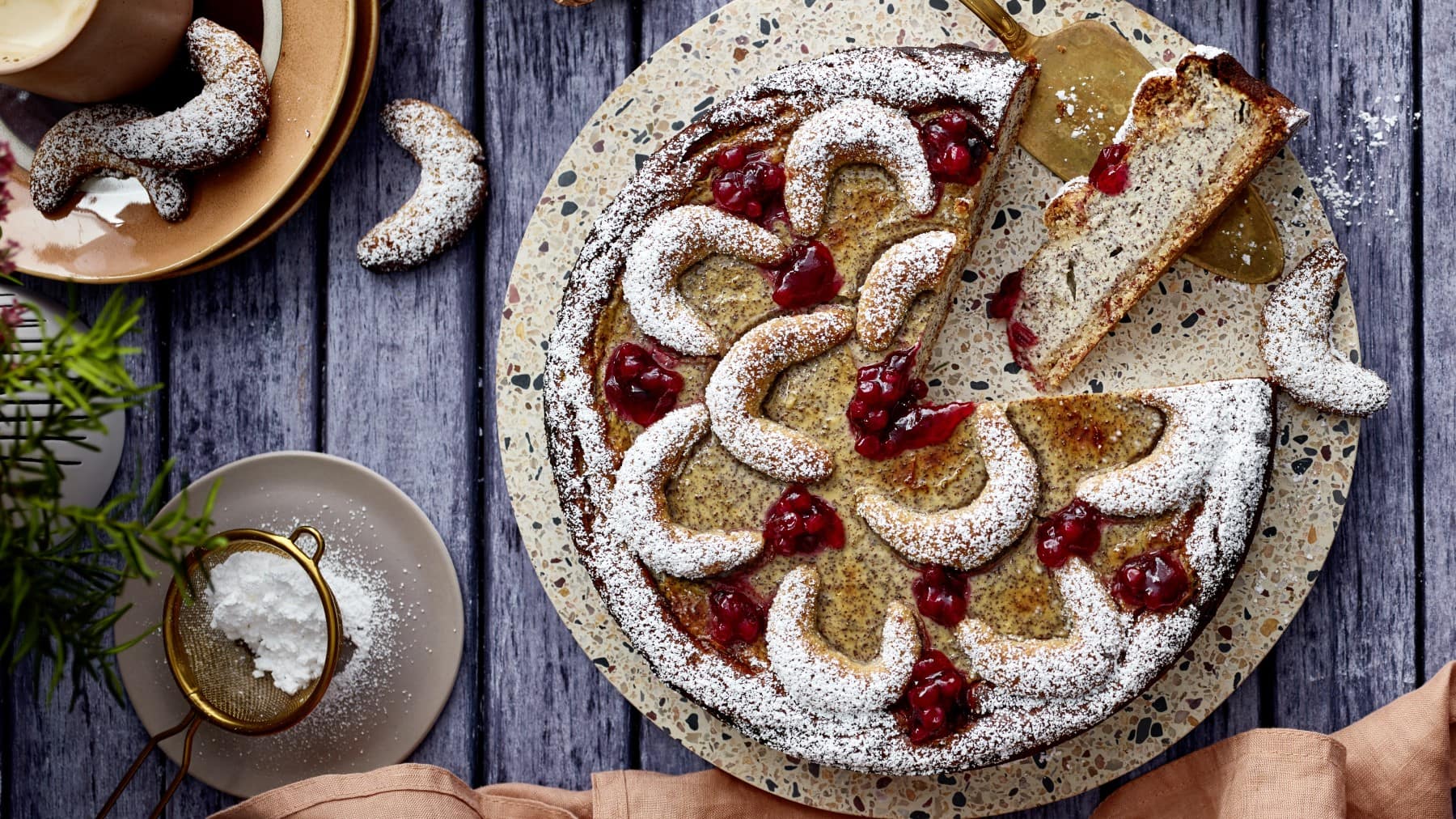 Ein Vanillekipferl-Kuchen auf einem Mosaik-Servierteller auf einem hölzernen Hintergrund. Links ein Teller mit Vanillekipferl und links unten ein kleinen Sieb mit Puderzucker.