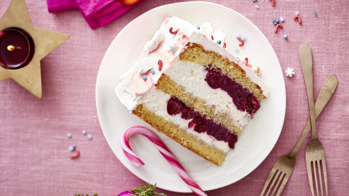Ein Stück Zuckerstangen-Torte auf einem weißen Teller auf einem rosa Hintergrund. Rechts auf dem Teller zwei Dessertgabeln. Links ein Teelicht.