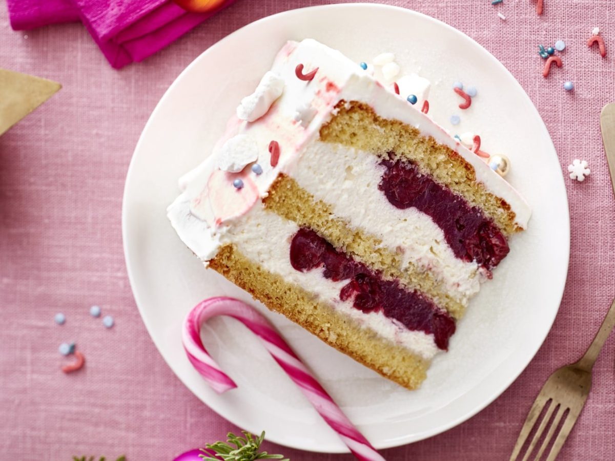 Ein Stück Zuckerstangen-Torte auf einem weißen Teller auf einem rosa Hintergrund. Rechts auf dem Teller zwei Dessertgabeln. Links ein Teelicht.