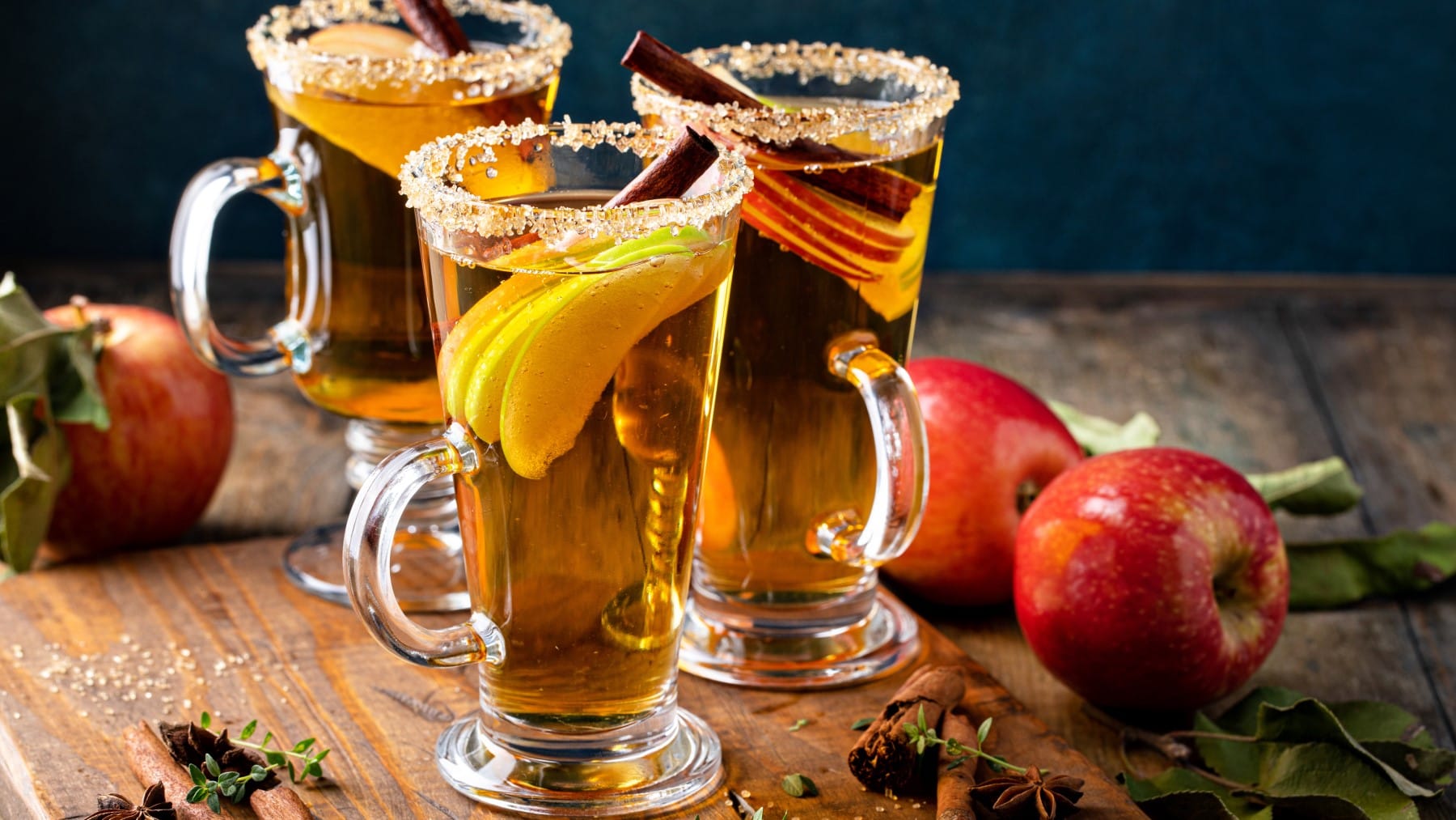 Drei Gläser Apfel Hot Toddy mit Apfelscheiben und Zuckerrand auf Holzuntergrund, drum herum Äpfel und Gewürze.