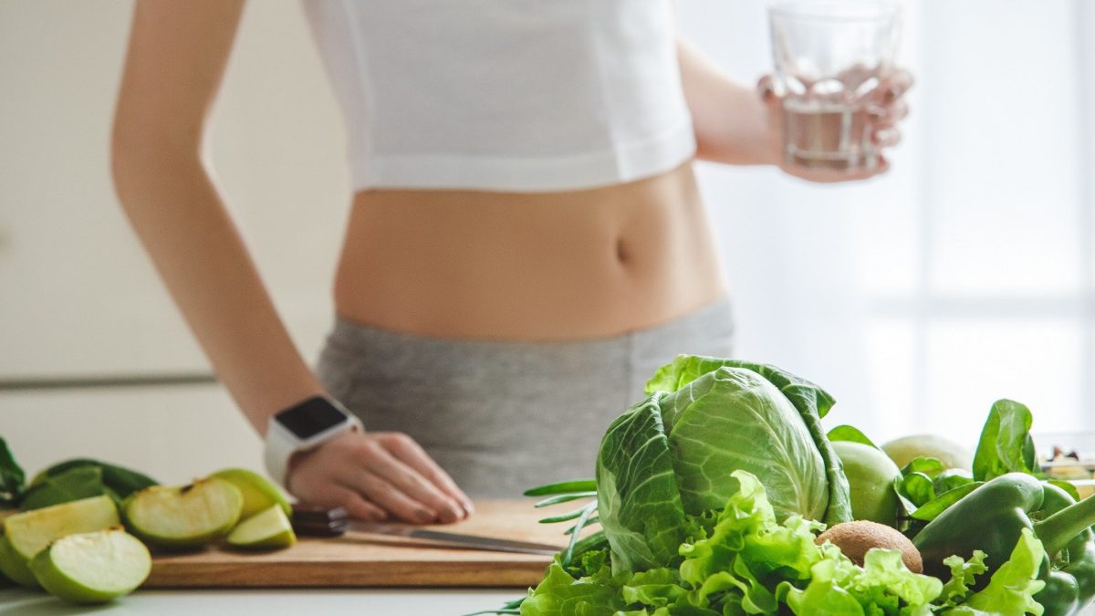 Frontal: eine schlanke Frau in einer Küche. Vor ihr liegen gesunde Detox-Lebensmittel, wie etwa Salat.