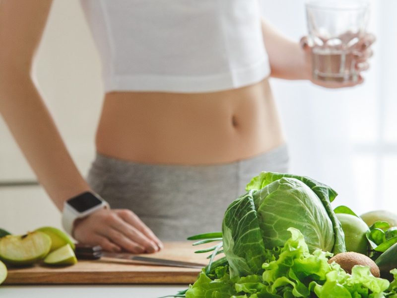 Frontal: eine schlanke Frau in einer Küche. Vor ihr liegen gesunde Detox-Lebensmittel, wie etwa Salat.