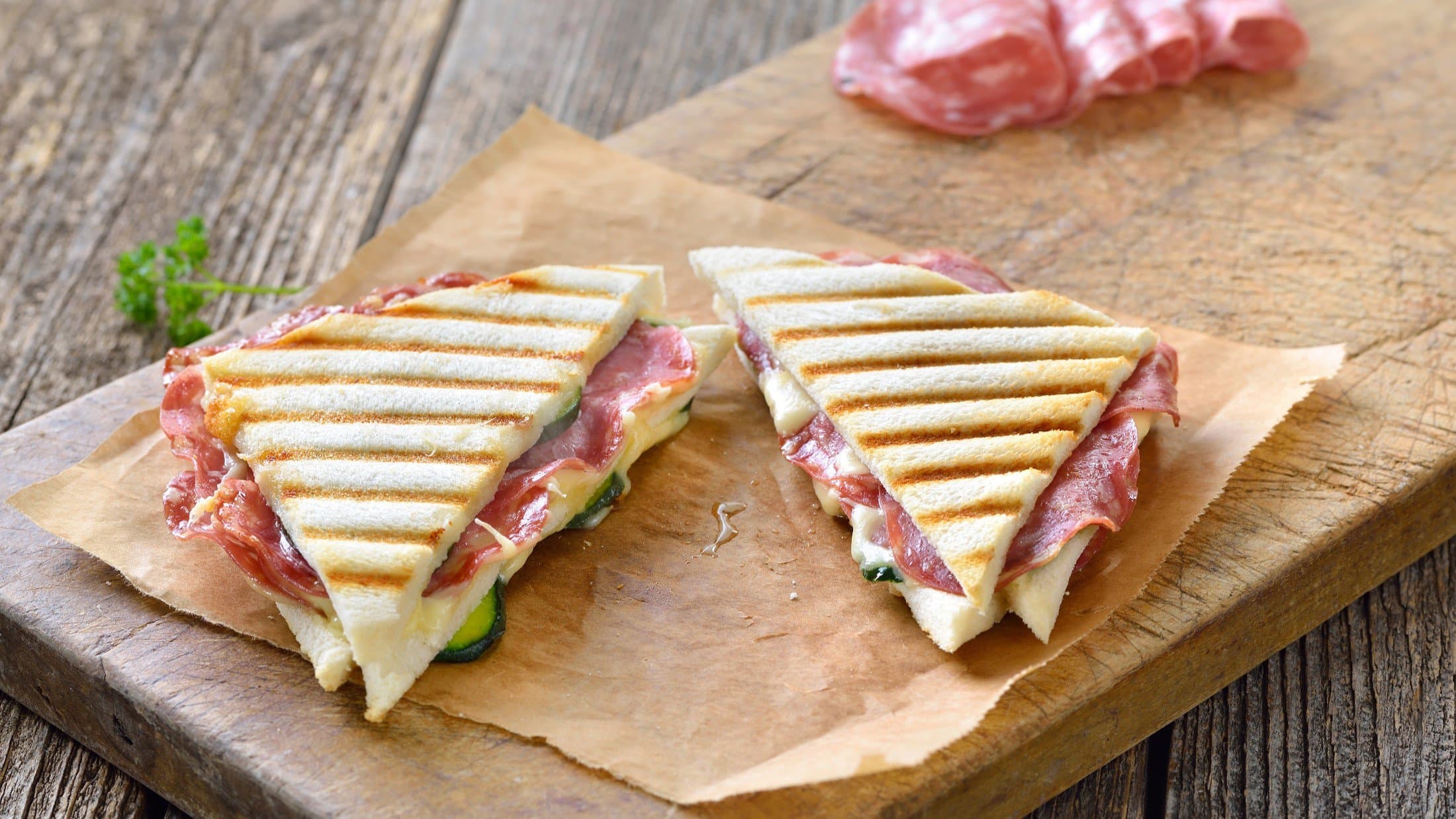 Draufsicht, schräg: Zwei italienische gegrillte Panini mit Salami und Käse liegen auf einem Stück Packpapier, Im Hintergrund ist aufgeschnittene Fenchelsalami.