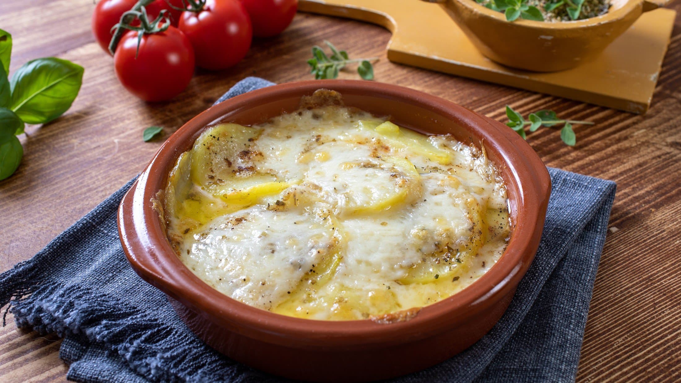 Draufsicht, schräg: Mozzarella-Kartoffeln aus dem Ofen: in einer ofenfestenForm stehen auf einem Holztisch. Drum herum sind Kräuter und Tomaten.