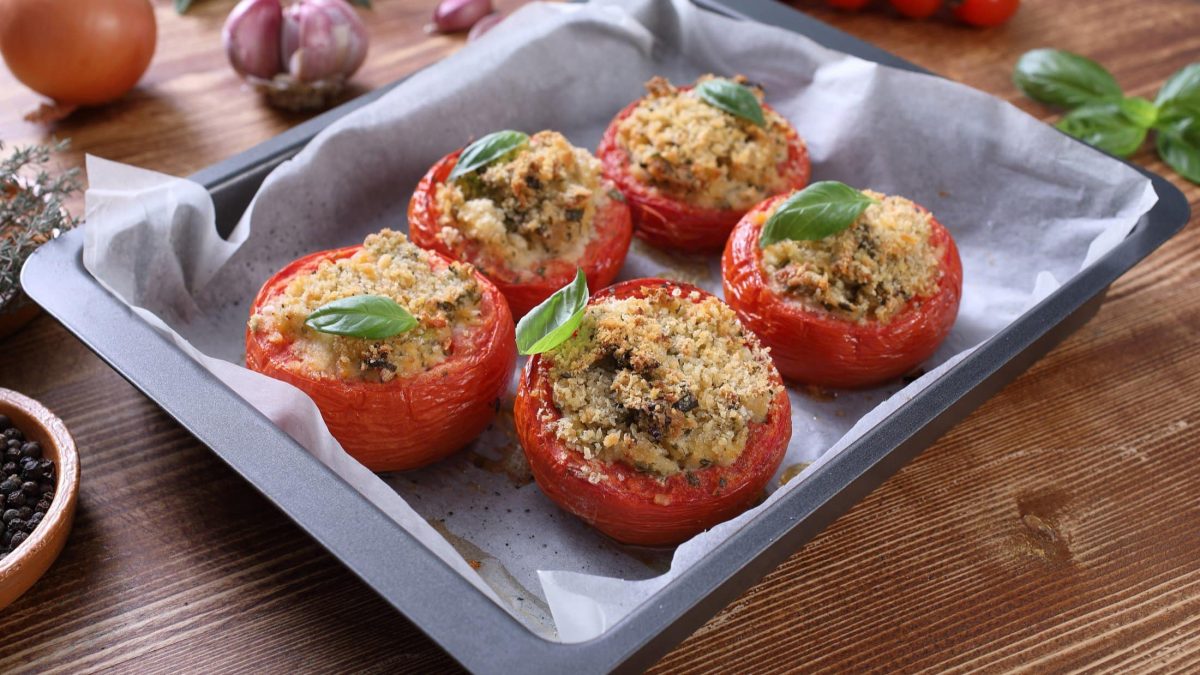 Draufsicht schräg: Ein Blech mit gefüllten Tomaten italienischer Art. Drum herum liegen Gewürze und Zutaten.