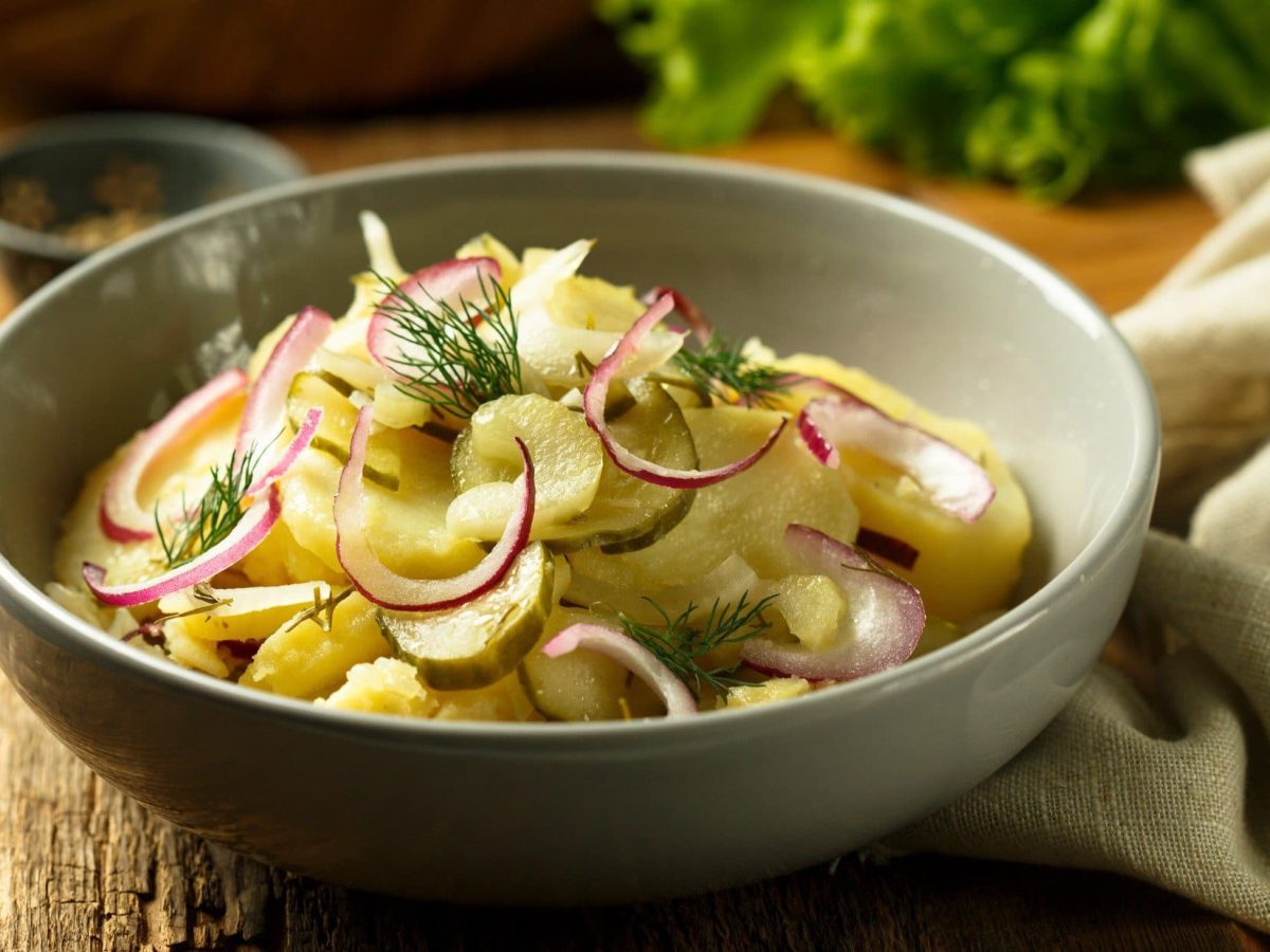 Draufsicht, schräg: Eine runde, graue Schüssel Kartoffelsalat mit Gewürzgurken steht auf einem Holztisch. Drum herum liegen ein Tuch und Gemüse.