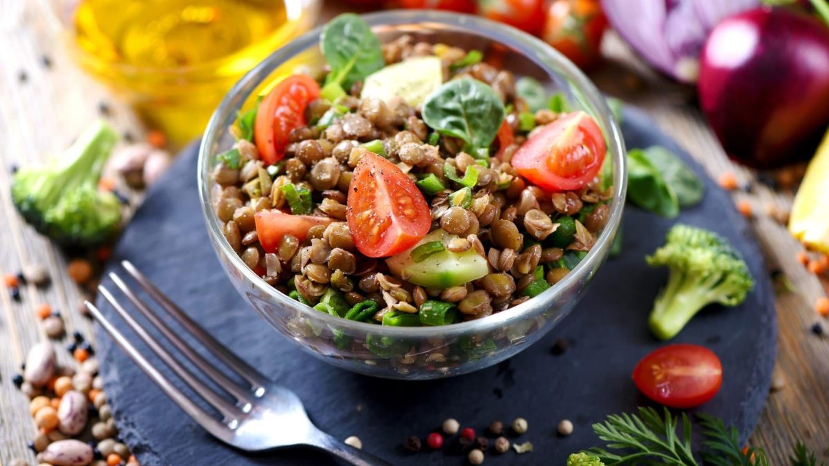 Draufsicht schräg: Ein Linsensalat mit Spinat und frischem Gemüse in einer Salatschüssel aus Glas. Drum herum liegt noch mehr Gemüse, Gewürze sowie eine Karaffe mit Öl.