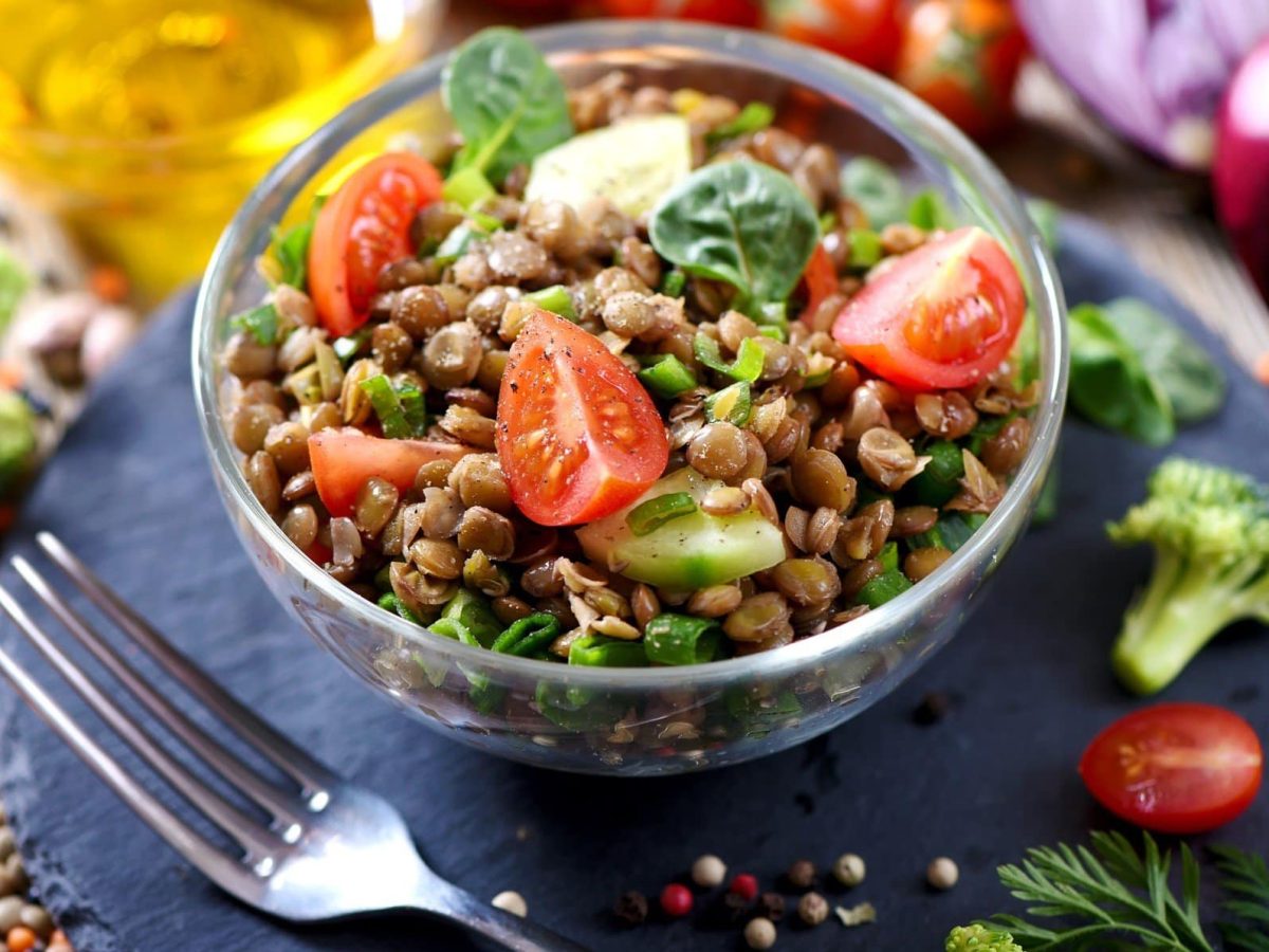 Draufsicht schräg: Ein Linsensalat mit Spinat und frischem Gemüse in einer Salatschüssel aus Glas. Drum herum liegt noch mehr Gemüse, Gewürze sowie eine Karaffe mit Öl.