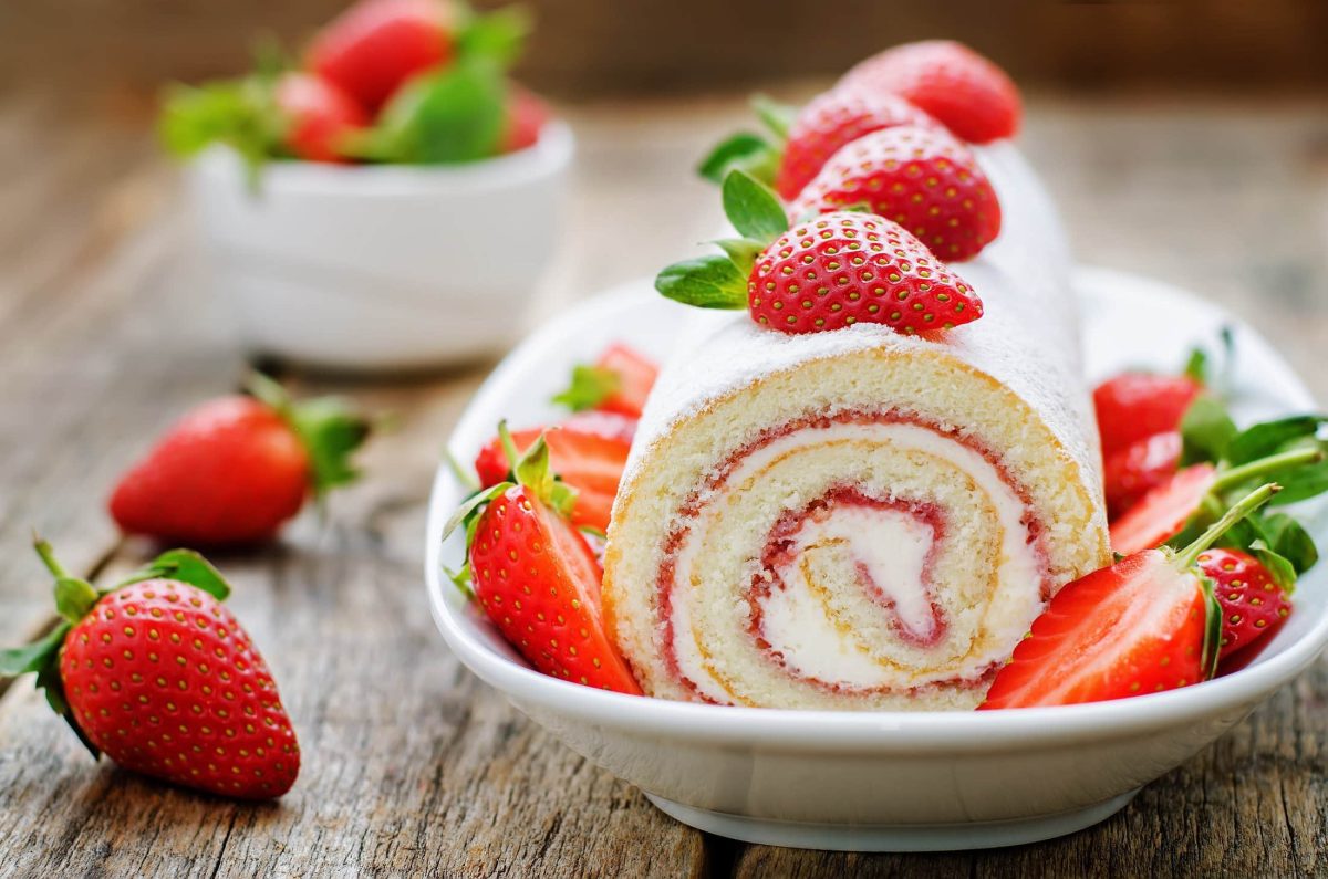 Frontal: eine angeschnittene Erdbeer-Roll aus Biskuit auf einem Teller, der auf einem Untergrund aus Holz steht. Die Erdbeer-Rolle ist mit unserem Profi-Tipp für perfekten Biskuit besonders gelungen.