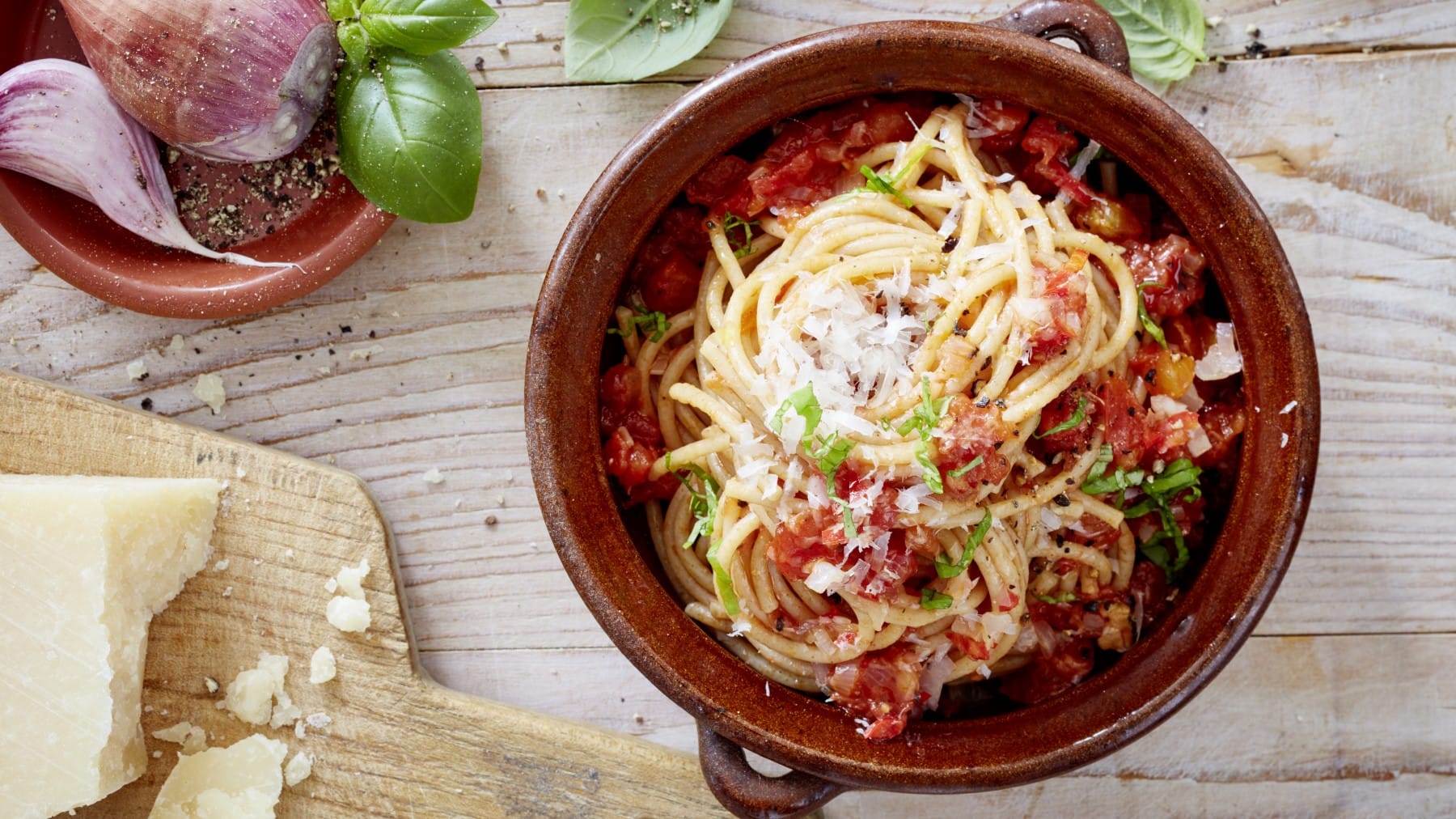 Spaghetti mit Chili in einem braunen Topf auf hölzernem Hintergrund. Links davon ein Brett mit Parmesan und links oben Schalotten, Knoblauch, Basilikum und Pfeffer.