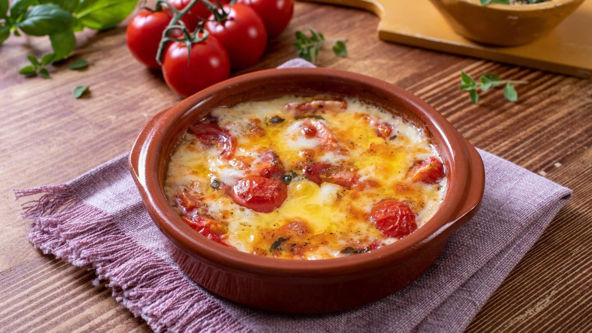Draufsicht schräg: überbackene Tomaten mit Käse in einer runden ofenfesten Form liegt auf einem karierten Küchentuch. Im Hintergrund liegen frische Tomaten und frisches Basilikum.