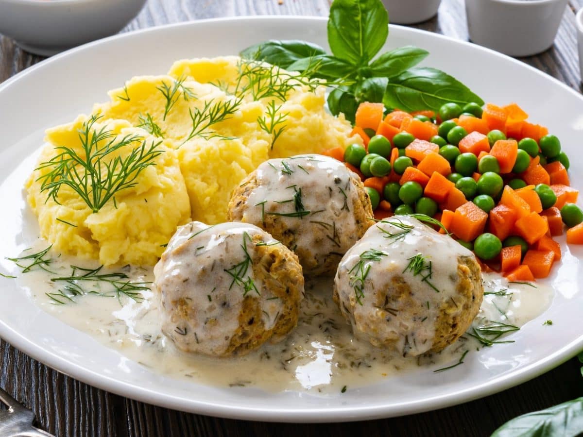 Draufsicht, schräg: Ein weißer Teller mit Fleischbällchen in Dillsauce sowie Kartoffelpüree und Möhren-Erbsen-Gemüse steht auf einem Holztisch.