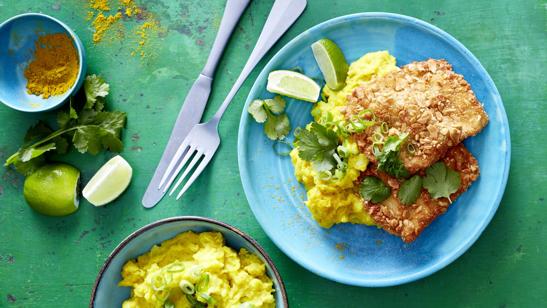 Erdnuss-Schnitzel mit Curry-Stampf auf einem blauen Teller auf grünem Hintergrund. Links davon Messer, Gabel, Curry und Limette. Unten auf dem Bild eine Schüssel mit Kartoffelstampf.
