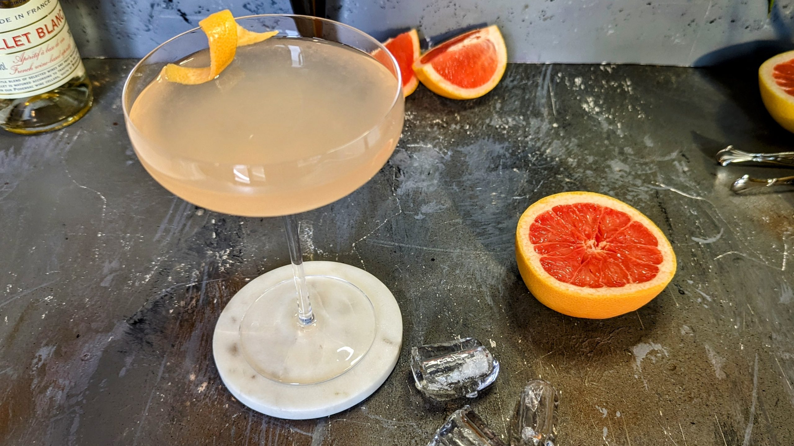 Eine Cocktailschale mit dem Drink auf Steinuntergrund von schräg oben fotografiert. Daneben Grapefruit und Cocktailzubehör.