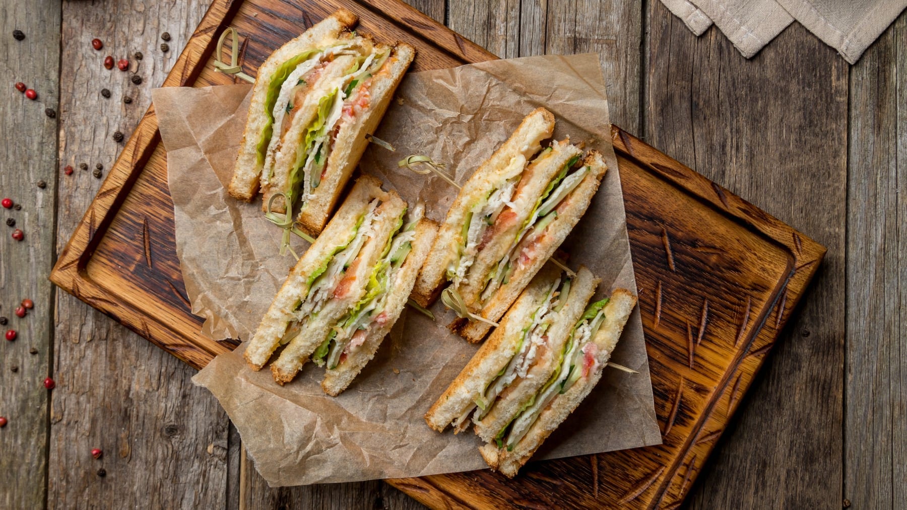 Veganes Club-Sandwich auf Backpapier auf einem Schneidebrett. Der Hintergrund ist hölzern und Pfefferkörner darauf verteilt.