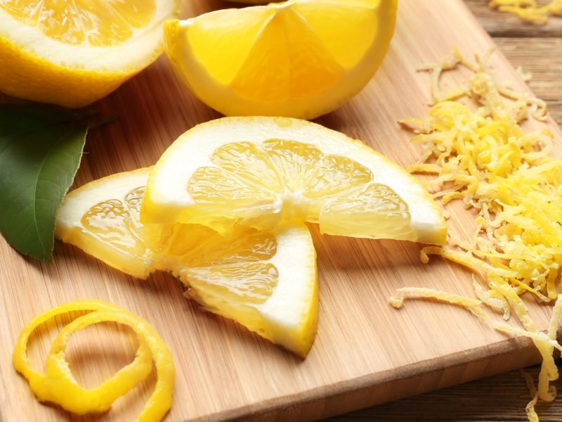 Draufsicht schräg: Auf einem Holzbrettchen liegen frische Zitronenstücke, Zitronenscheiben und Zesten. Es gibt eine breite Zeste und mehrere dünne.
