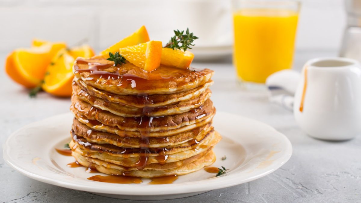 Zitronen-Pancakes mit Orangensirup auf einen Teller gestapelt mit Orangenstücken darauf und daneben, heller Hintergrund.