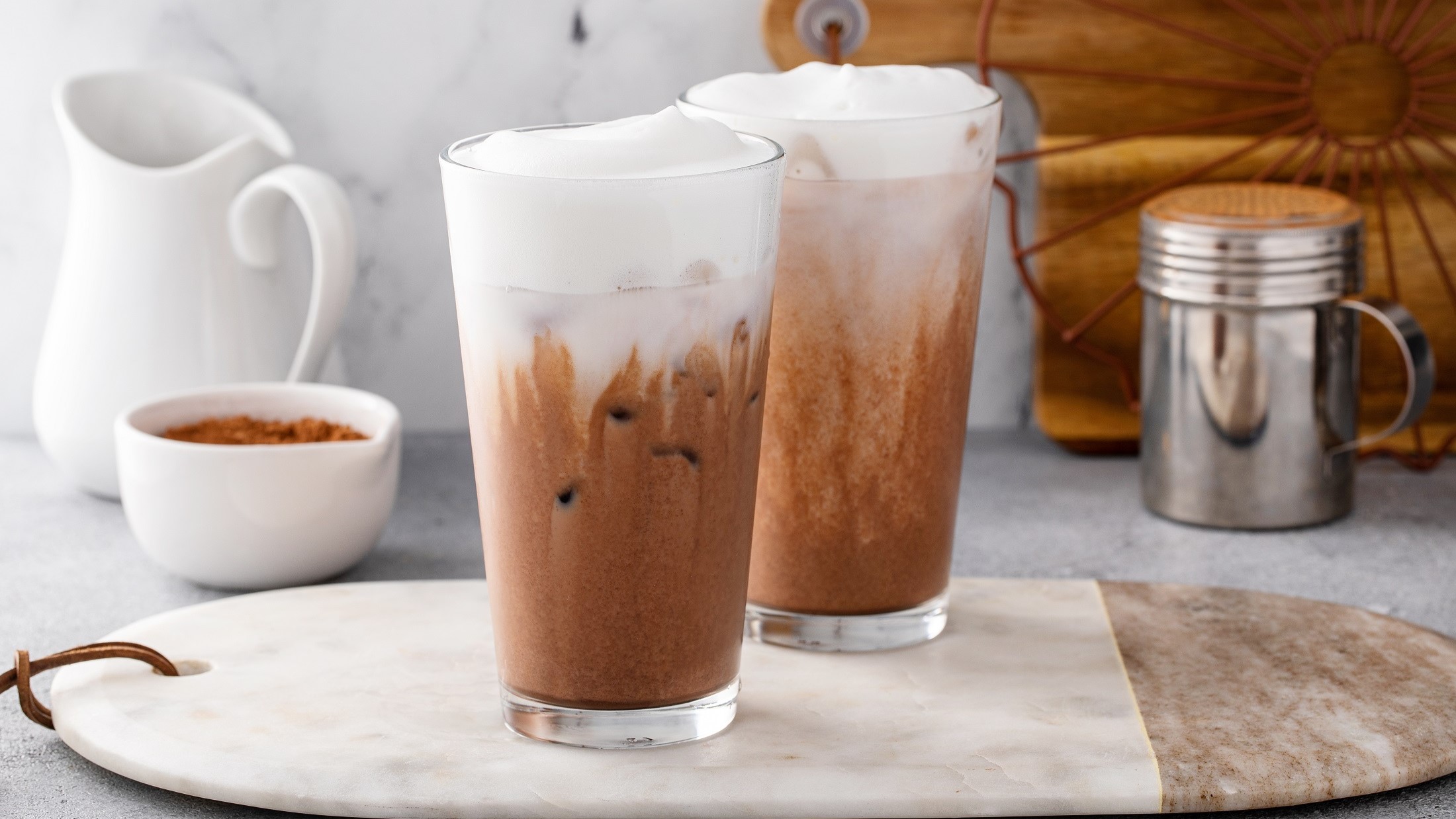 Frontal: Zwei Gläser Chocolate-Chai stehen auf einem Holzbrettchen. Das liegt auf einem weißen Untergrund aus Marmor. Naneben stehen eine weiße Milchkanne aus Porzellan, eine Tasse mit braunem Zucker und ein Kakao-Streuer aus Edelstahl.