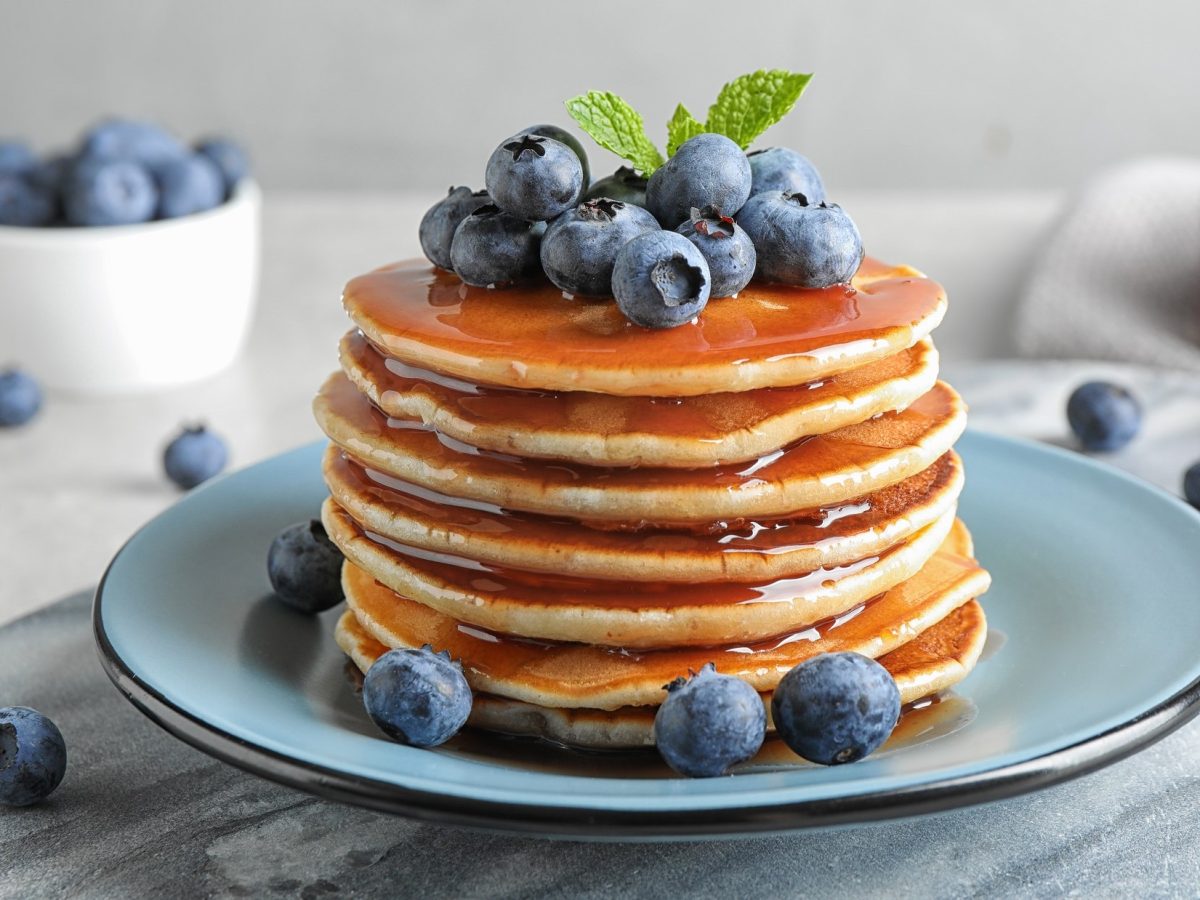 Frontal: Auf einem Tisch steht ein Bläulicher Teller mit einem Turm Dinkel-Joghurt-Pancakes. Auf den Pancakes sind Blaubeeren. Die Blaubeeren sind auch auf dem Tisch verteilt.