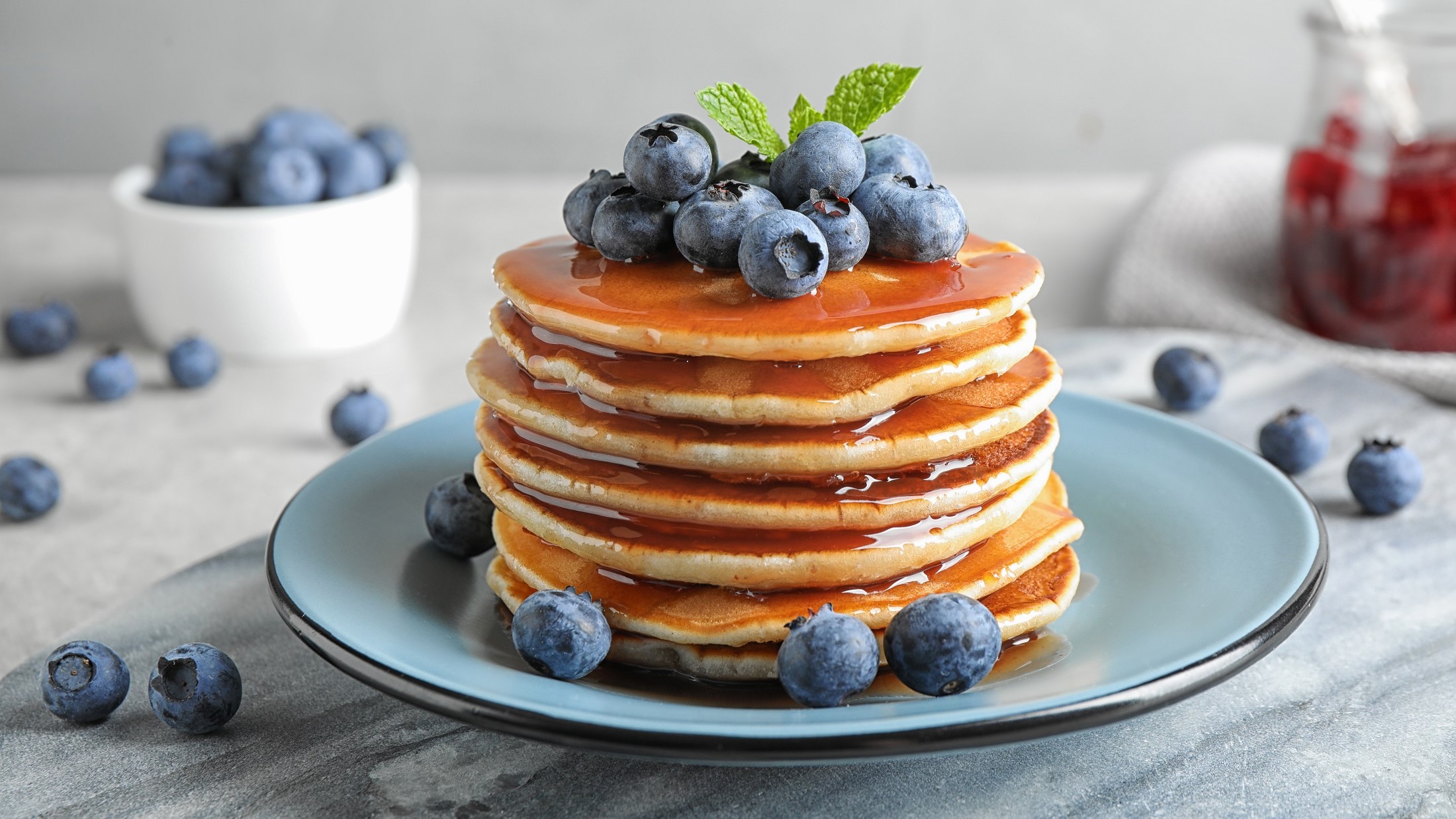 Frontal: Auf einem Tisch steht ein Bläulicher Teller mit einem Turm Dinkel-Joghurt-Pancakes. Auf den Pancakes sind Blaubeeren. Die Blaubeeren sind auch auf dem Tisch verteilt.