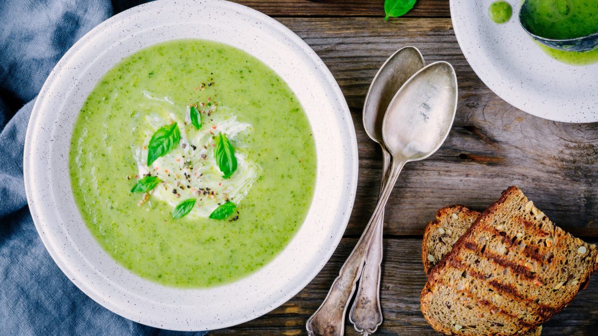 Draufsicht: ein weißer Teller mit einer Frühlingskräuter-Suppe mit Spinat steht auf einem Tisch. Daneben liegen zwei geröstete Brotscheiben und zwei Löffel.