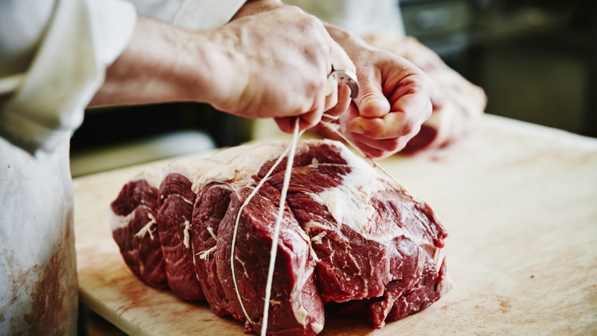 Frontal: Zwei Hände eines Mannes knoten einen großes Stück rohes Tierfleisch mit einem Bindfaden zusammen. Das Fleisch liegt auf einem Holzbrettchen. Das Foto macht es leicht, gutes Fleisch erkennen.