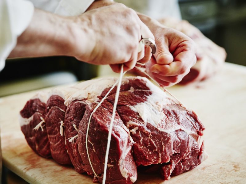 Frontal: Zwei Hände eines Mannes knoten einen großes Stück rohes Tierfleisch mit einem Bindfaden zusammen. Das Fleisch liegt auf einem Holzbrettchen. Das Foto macht es leicht, gutes Fleisch erkennen.