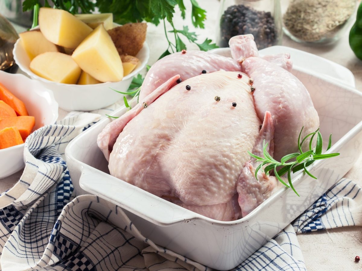 Draufsicht, schräg: Ein rohes Hähnchen liegt in einer Auflaufform. Drum herum stehen weiße kleine Schüsseln mit geschnittenem Gemüse, Kräuter, Öl und Gewürzen. Aus ihnen soll ein Ofenhähnchen mit Gemüse zubereitet werden.