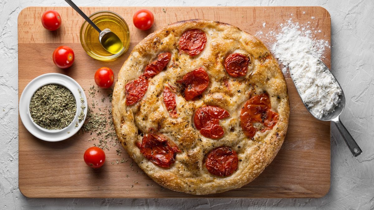 Focaccia barese mit Tomaten auf einem Holzbrett mit Olivenöl, Oregano, Mehl und frischen Tomaten.