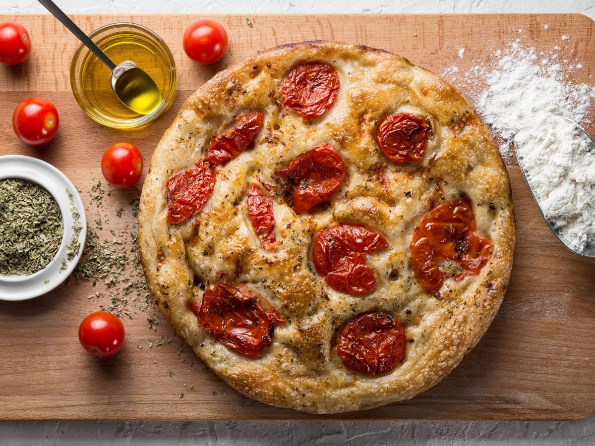 Focaccia barese mit Tomaten auf einem Holzbrett mit Olivenöl, Oregano, Mehl und frischen Tomaten.